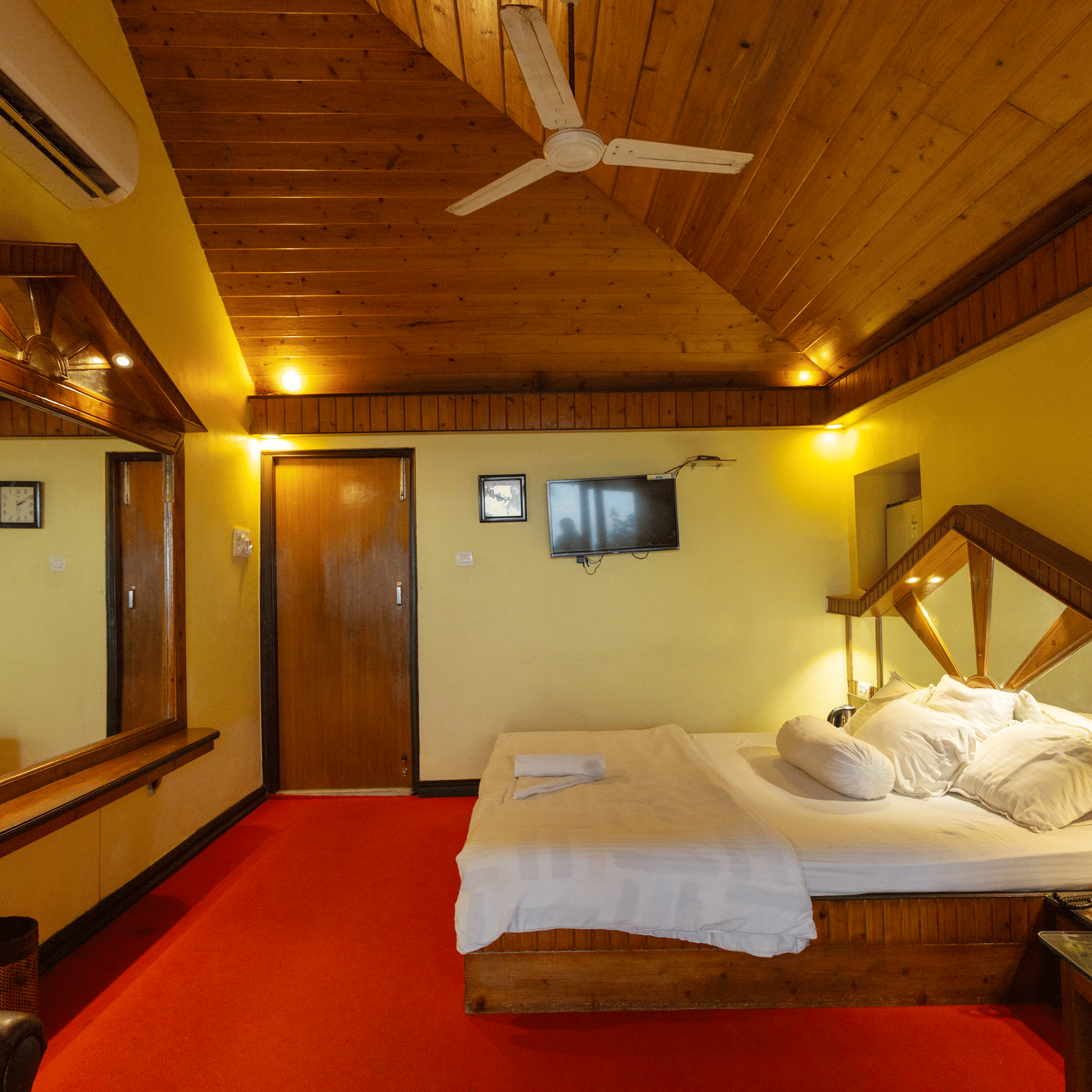 A bedroom with a king size bed, a wooden ceiling, a large mirror, a bedside table, and a wall mounted TV at Hotel Seagull Digha.